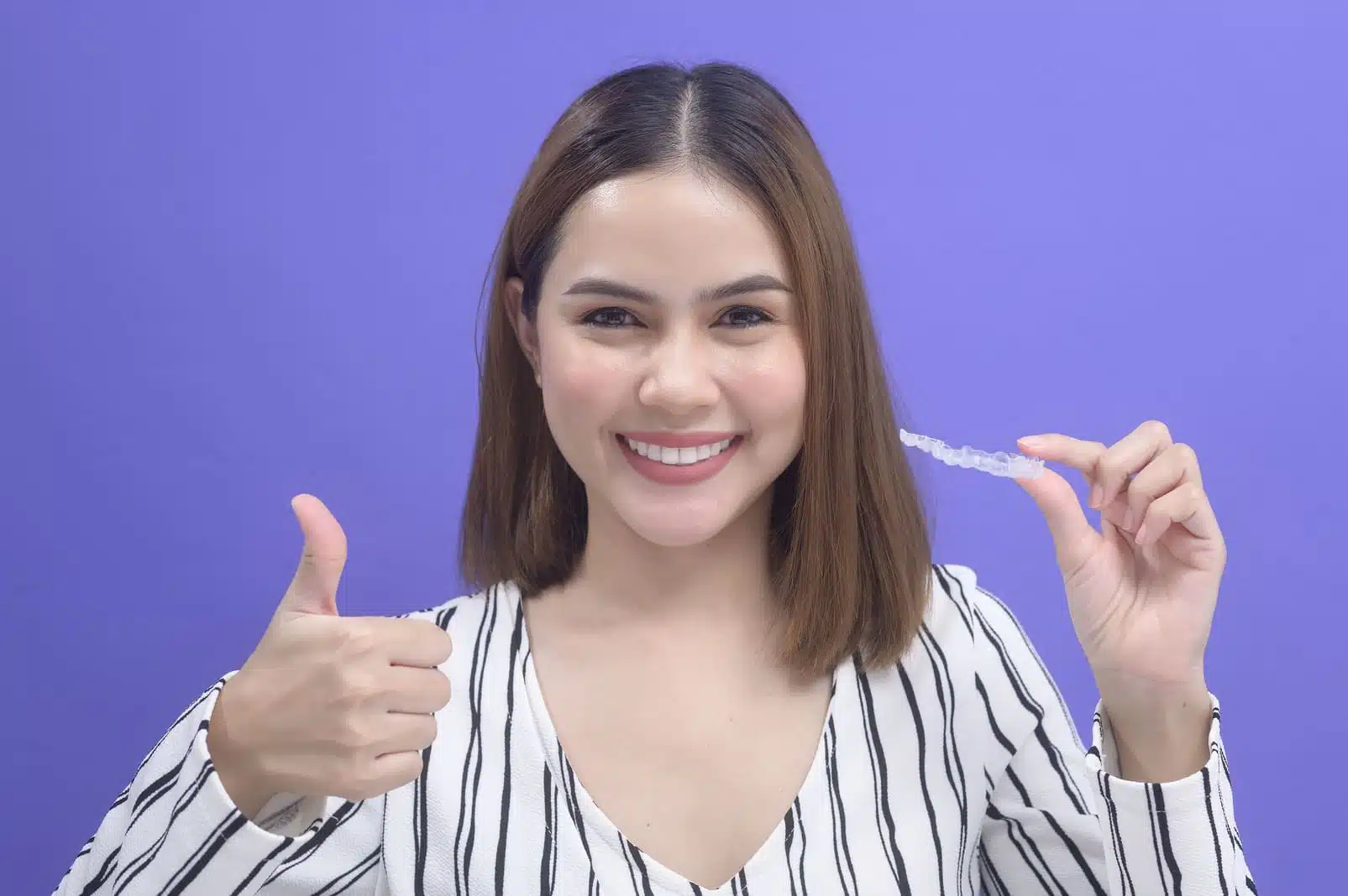 Smiling woman holding Invisalign clear aligner with a thumbs up, against a purple background, illustrating the comfort and effectiveness of Invisalign orthodontic treatment.