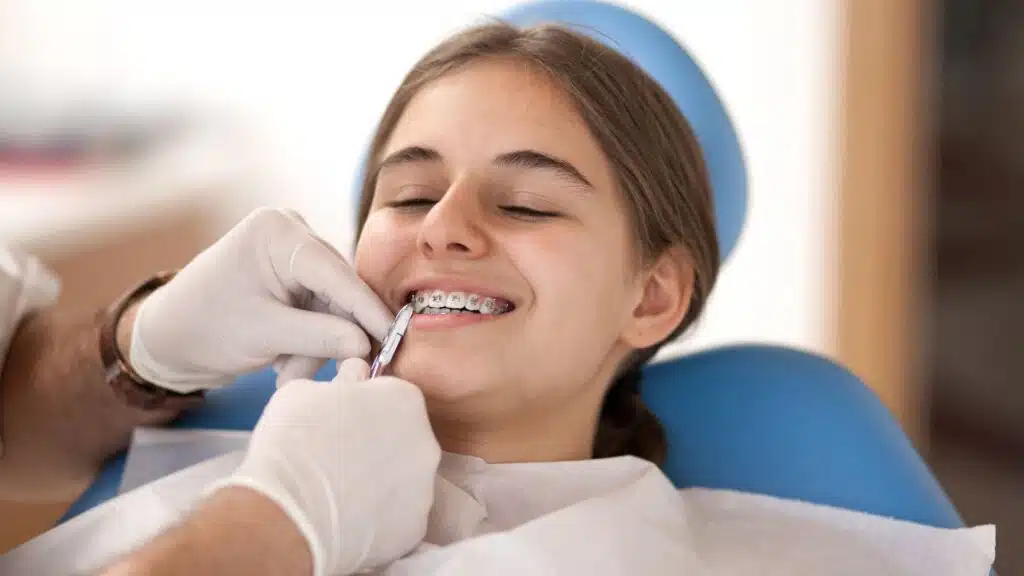 Teen girl with braces smiling in dental chair while orthodontist adjusts braces, related to braces cost and treatment options for kids and teens.