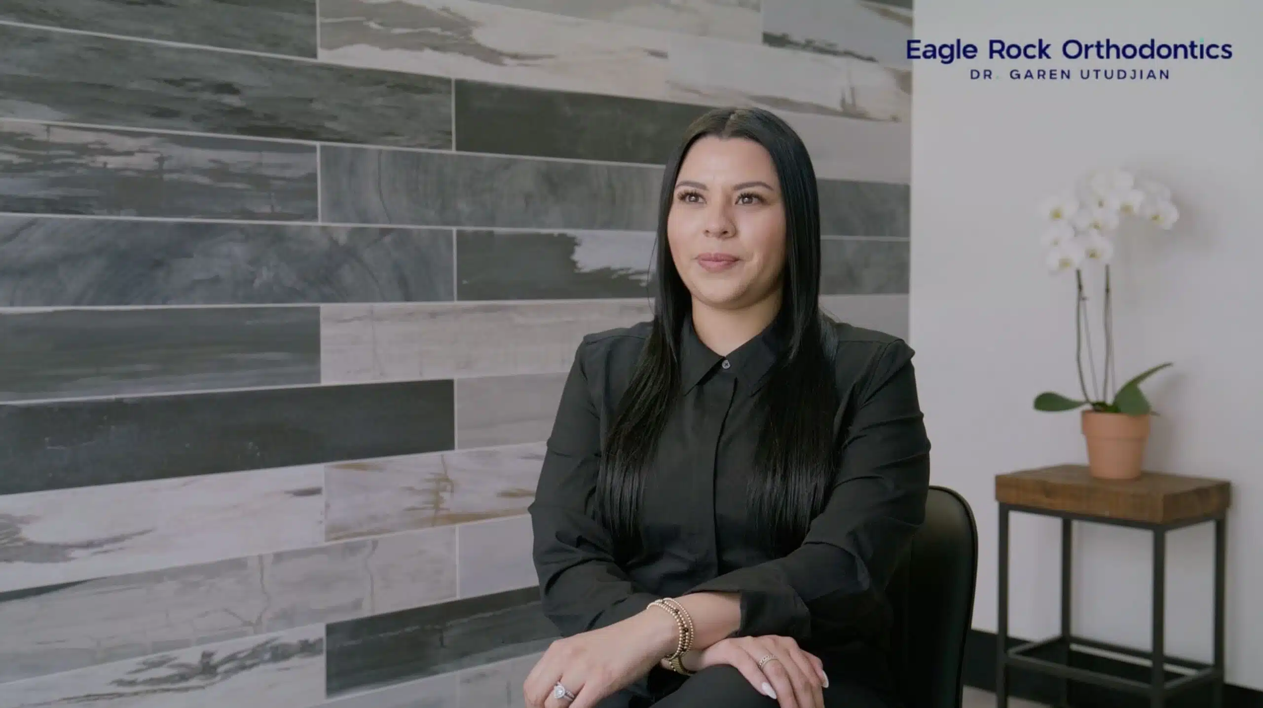 Patient testimonial at Eagle Rock Orthodontics featuring a woman in a black shirt, sitting in a modern office setting with a decorative wall and a potted plant.