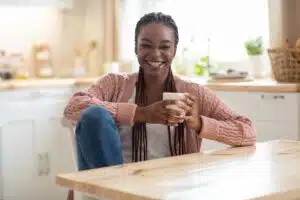 Smiling woman with braided hair sitting at a kitchen table, holding a cup, representing comfort and confidence in a relaxed home environment.