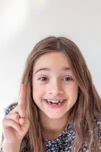 Smiling girl with a missing front tooth, raising one finger, representing the positive experience and comfort of pediatric orthodontic visits in Highland Park.