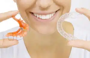 Person smiling and holding two types of retainers, one orange and one clear, emphasizing retainer options for maintaining dental alignment after orthodontic treatment.