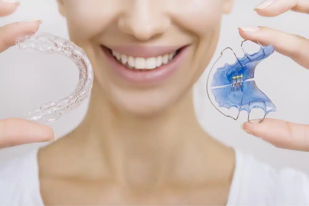 Woman smiling while holding a clear retainer and a blue orthodontic retainer, showcasing options for post-braces care.