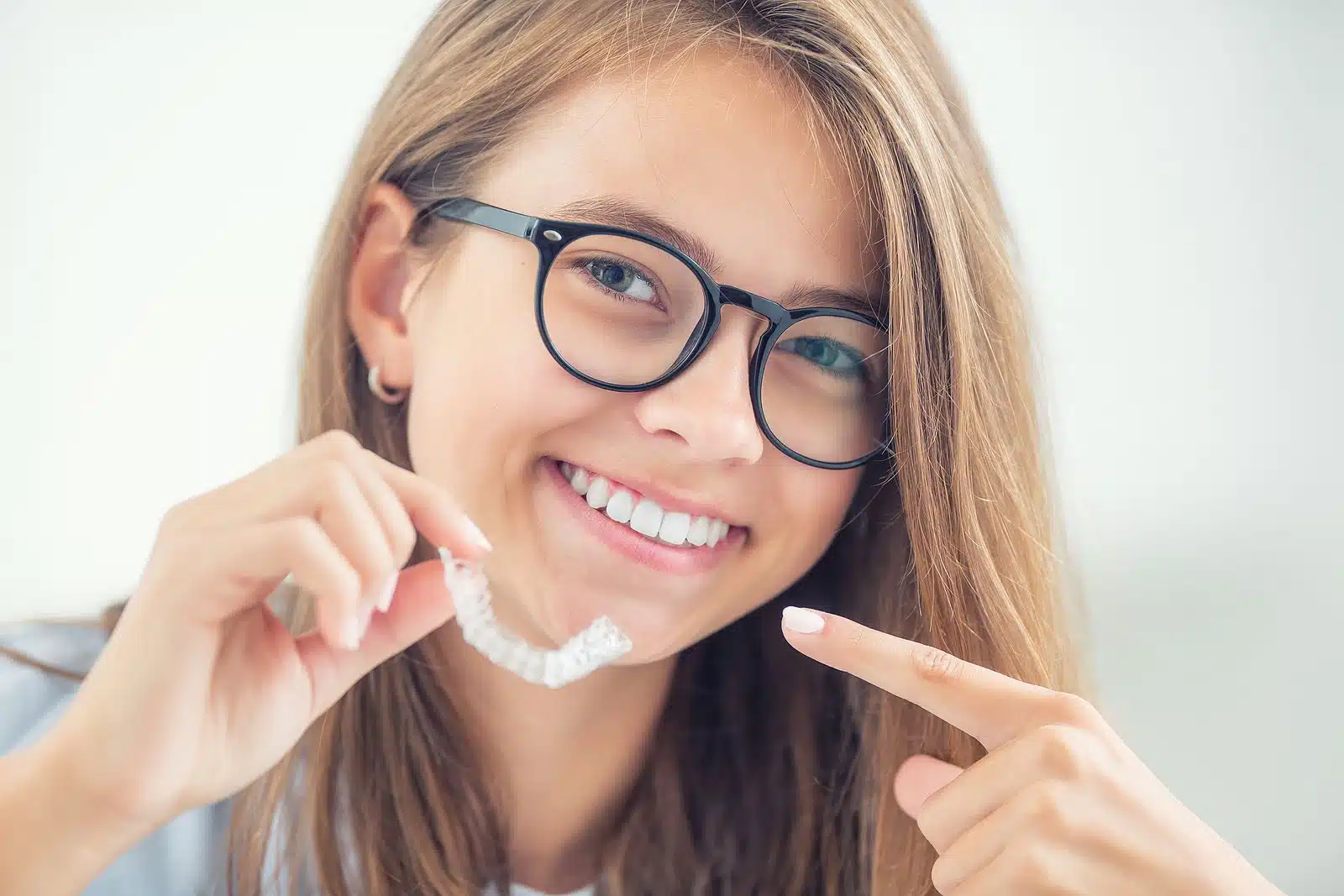 Smiling young woman wearing glasses holding clear aligner, promoting discreet teeth straightening with Invisalign.